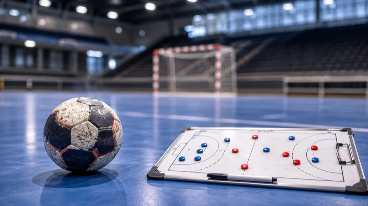 Ein Handball liegt auf dem Hallenboden neben einer Taktiktafel, im Hintergrund ist unscharf ein Handballtor in einer leeren Sporthalle zu sehen.